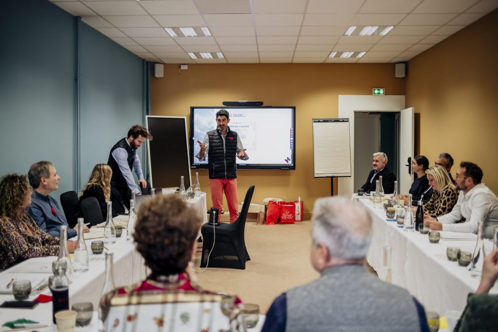 Photo d'une session de coaching d'équipe, l'équipe est placée tout autour des coachs avec une table en U et les regardent tout comme l'écran derrière eux.